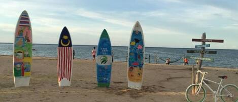 Tablas de surf con diversos diseños y una bicicleta en una playa arenosa, con un cartel de señalización al fondo.