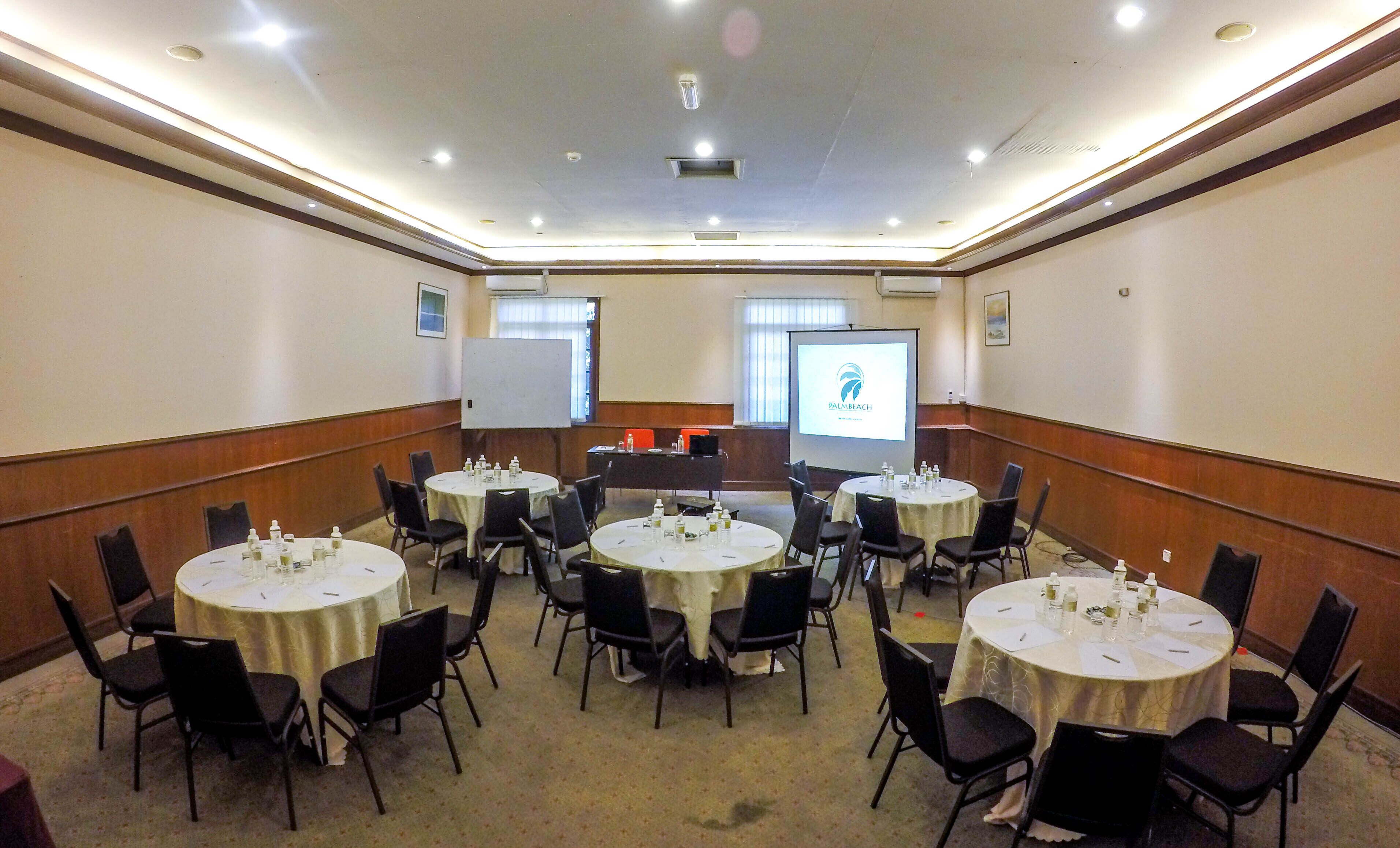 Un salón de conferencias con varias mesas redondas, sillas negras y una pantalla de proyector mostrando un logotipo.