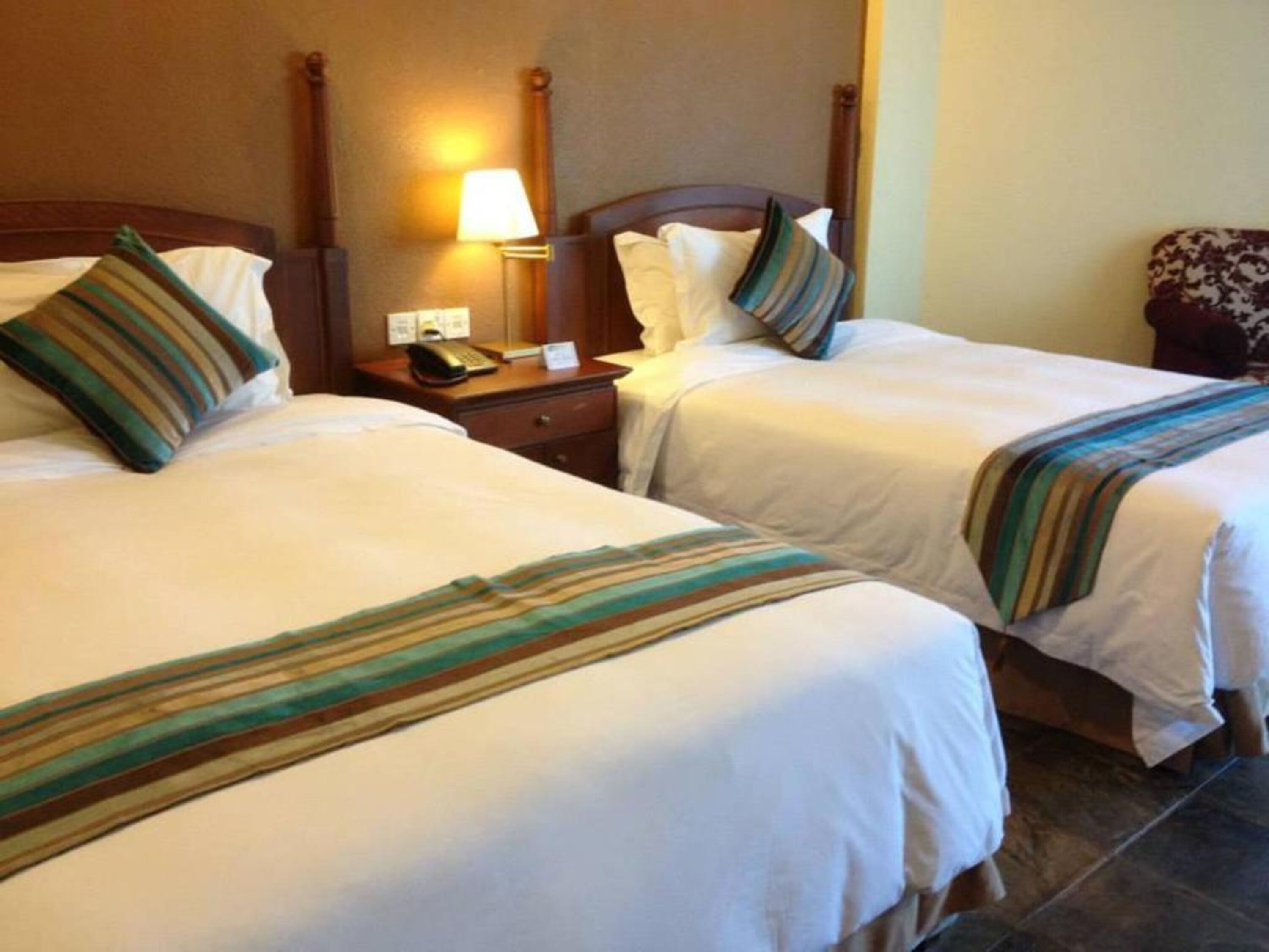 Habitación de hotel con dos camas individuales, cada una con una manta a rayas. Se aprecia un mesita de noche con una lámpara y un teléfono.