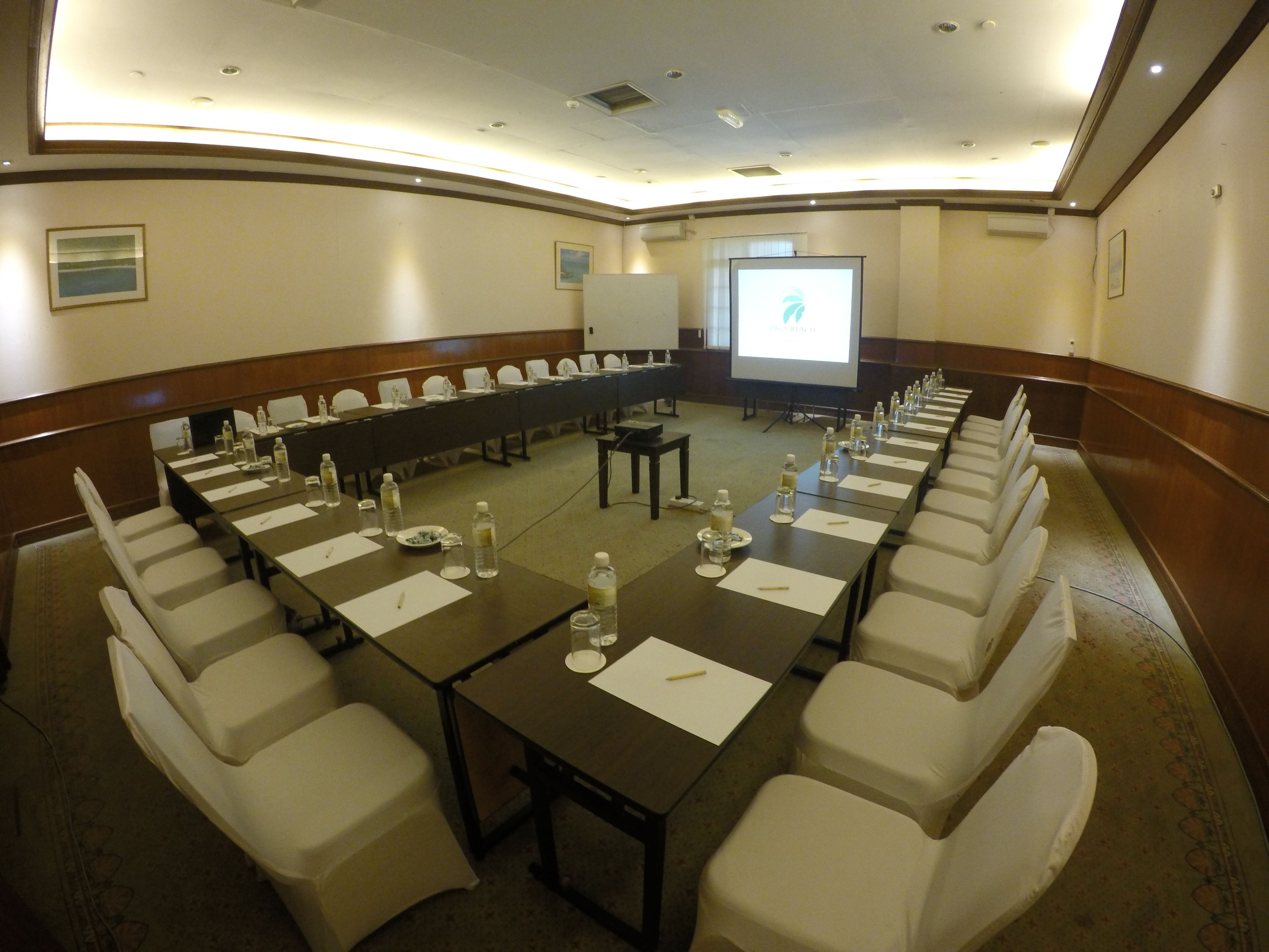 Un salón de conferencias con una larga mesa, sillas blancas, botellas de agua y una pantalla de proyector.
