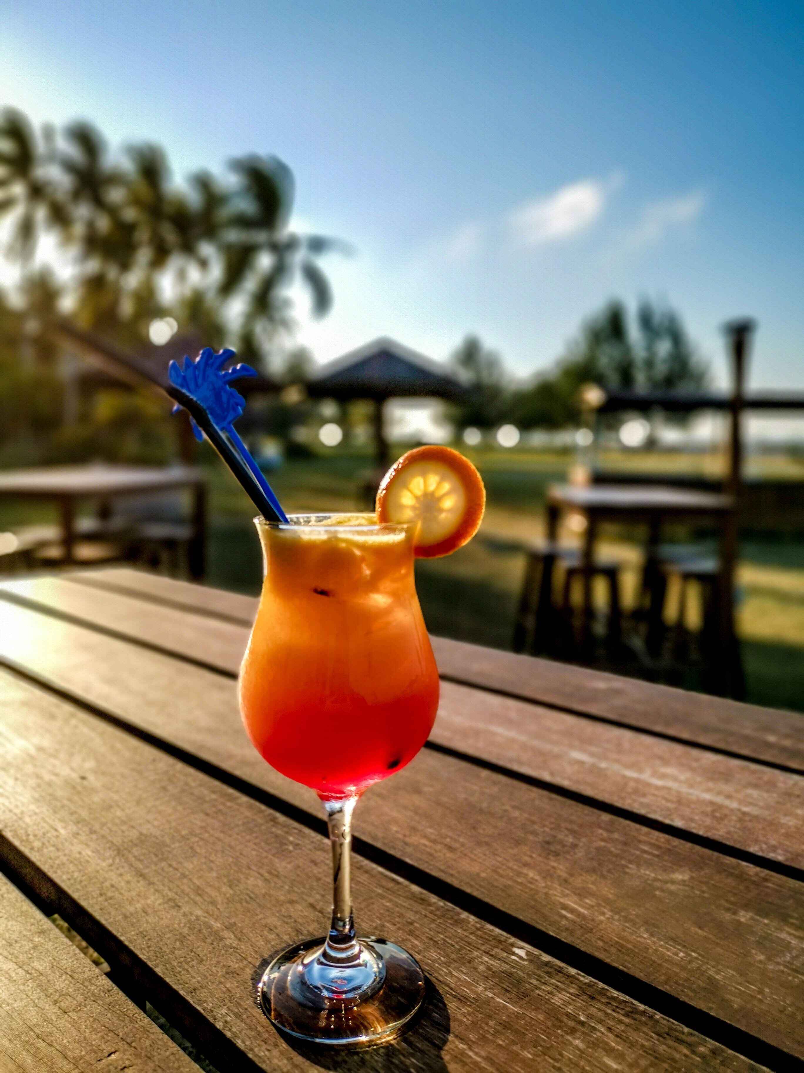 En cocktail i et glas med sugerør i blå farve og et appelsinstykke på kanten, stående på et træbord udendørs.