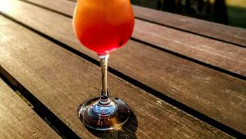En cocktail i et glas med sugerør i blå farve og et appelsinstykke på kanten, stående på et træbord udendørs.