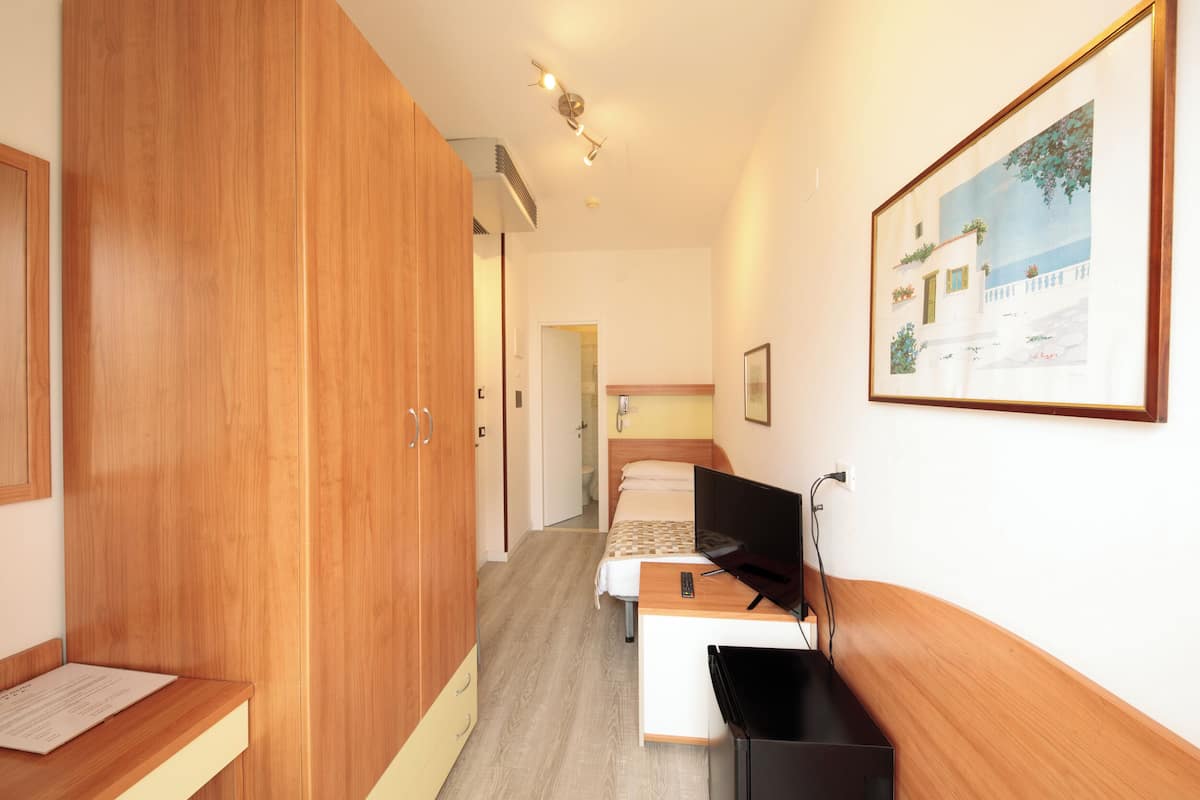Habitación de hotel con un armario de madera, una cama con cabecera blanca, un escritorio con televisor y un cuadro en la pared.
