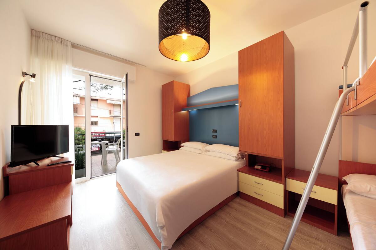 Habitación de hotel con cama, televisor, escritorio y balcón con vistas a edificios.