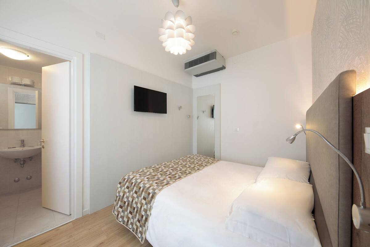 Un dormitorio con cama, mesita de noche, espejo y televisor instalado en la pared.