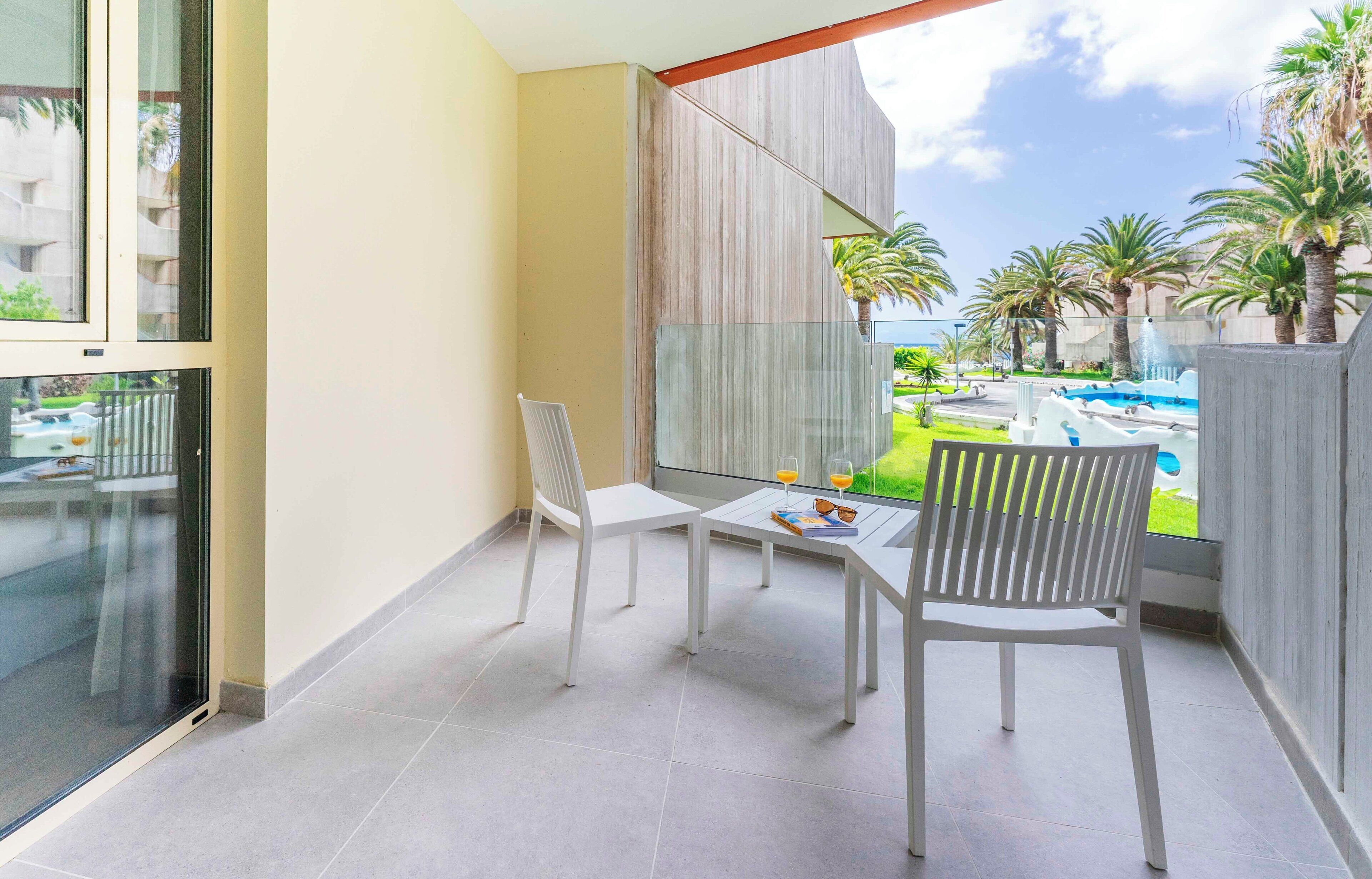 A balcony with a glass partition, white chairs, a small table, and a view of a swimming pool and palm trees.