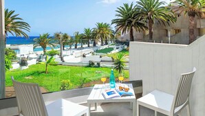 Ein Balkon mit weißen Stühlen, einem Tisch mit Getränken und einer Zeitschrift, und Blick auf einen Pool und Palmen.