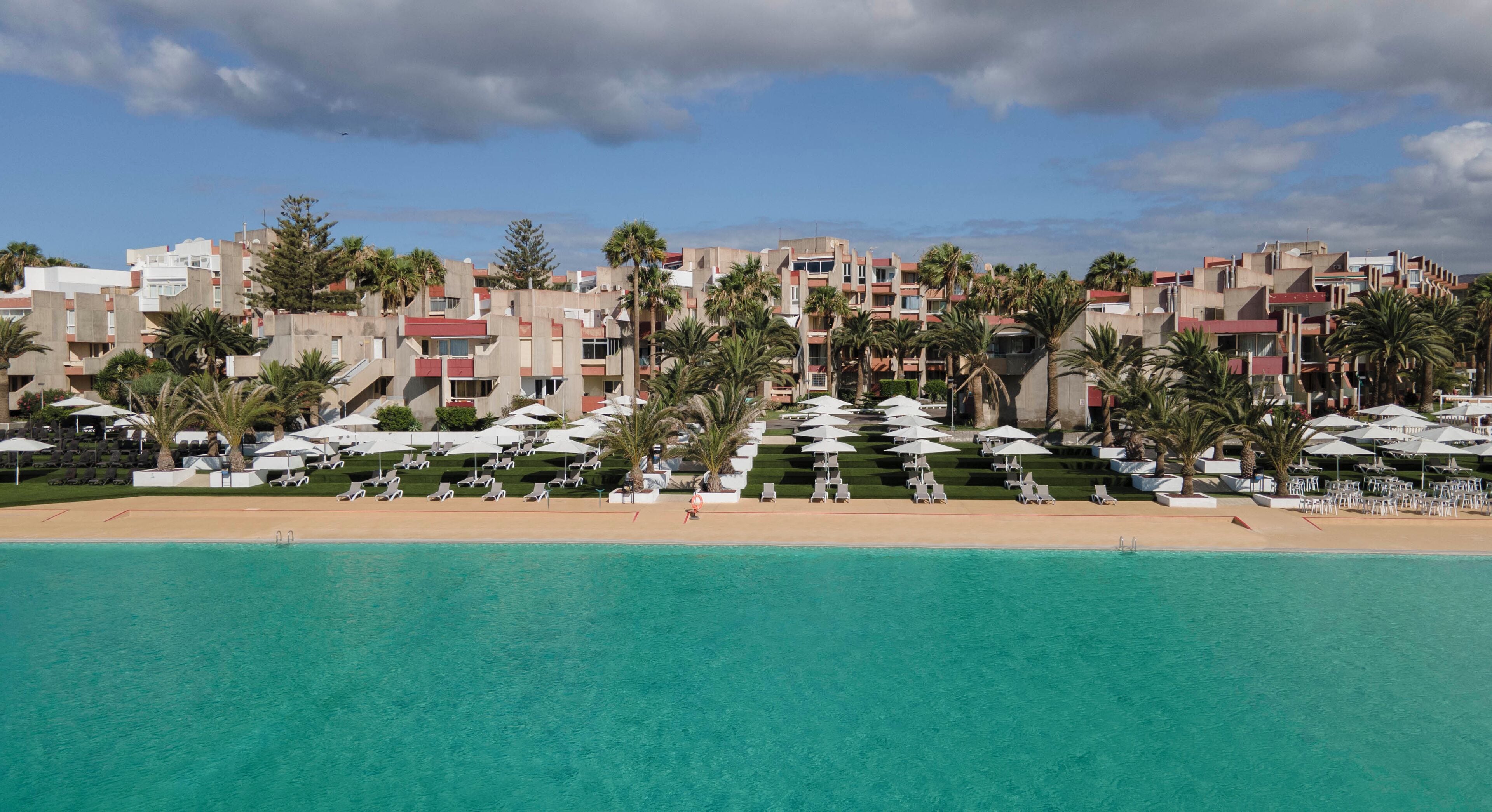 A beachfront hotel with multiple buildings, palm trees, and a clear blue sea.