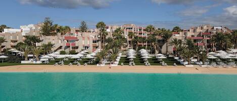 A beachfront hotel with multiple buildings, palm trees, and a clear blue sea.