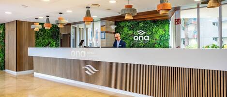A reception area with a wooden reception desk, a green wall with plants, and multiple pendant lights.