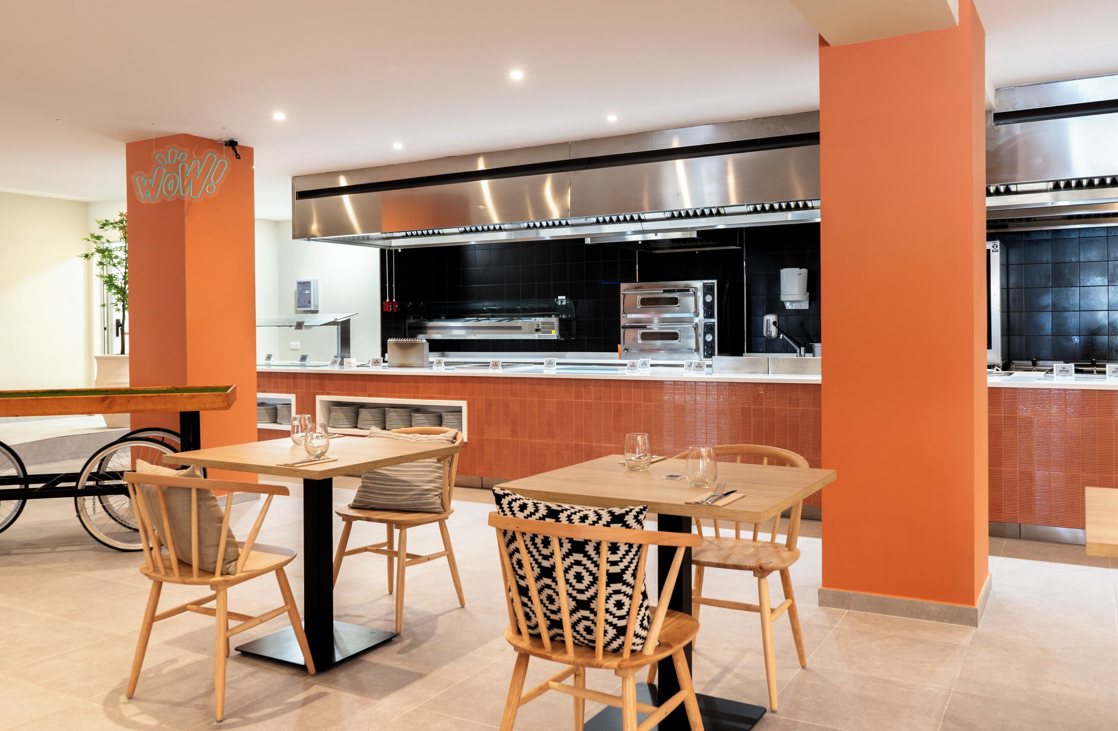 A modern restaurant with a central dining area featuring a long table and chairs, a kitchen with stainless steel appliances, and orange accent walls.