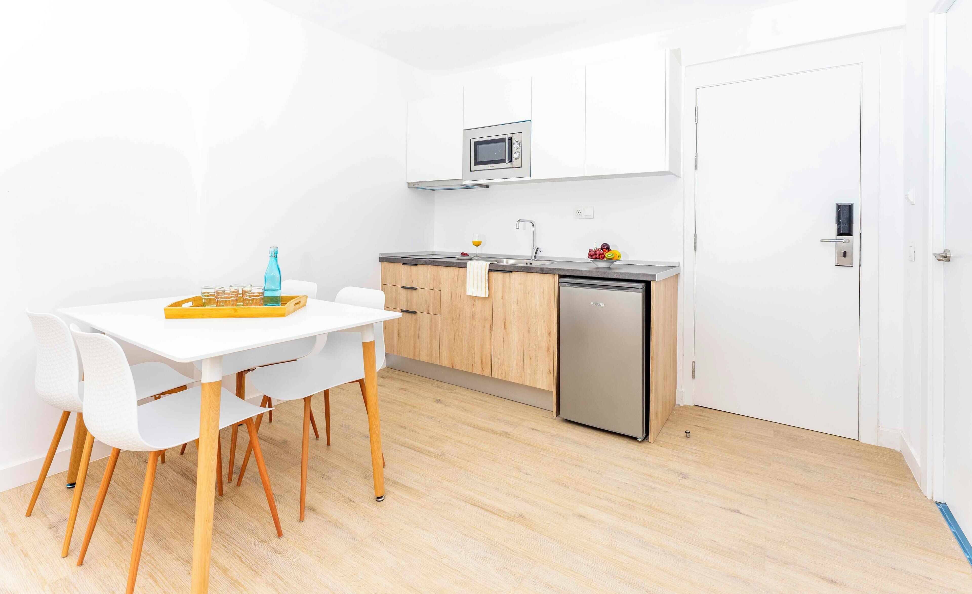 A compact kitchen with a microwave, sink, and dishwasher. There is a dining table with chairs and a tray with glasses and a bottle.