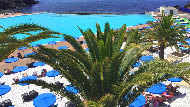 Ein Poolbereich mit blauen Sonnenschirmen und Liegestühlen mit Blick auf das klare, blaue Meer.