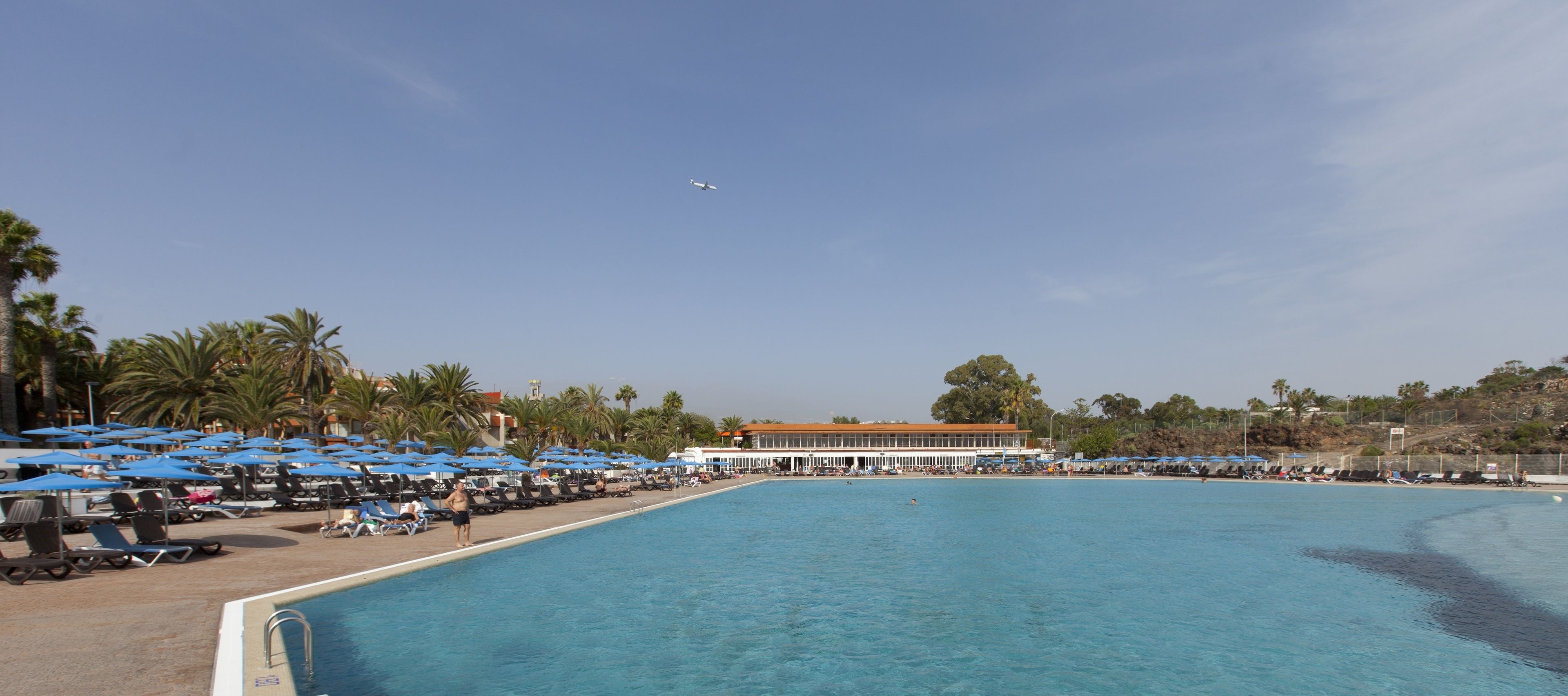 A large pool with lounge chairs and umbrellas, a building with a red roof, and palm trees in the background.