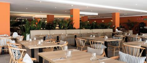 A modern restaurant with wooden tables and chairs, some with patterned cushions. There are potted plants and a seating area with a sofa.
