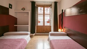 Habitación de hotel con dos camas individuales, cabecero de madera, balcón con vistas y una pequeña estantería con un jarrón.
