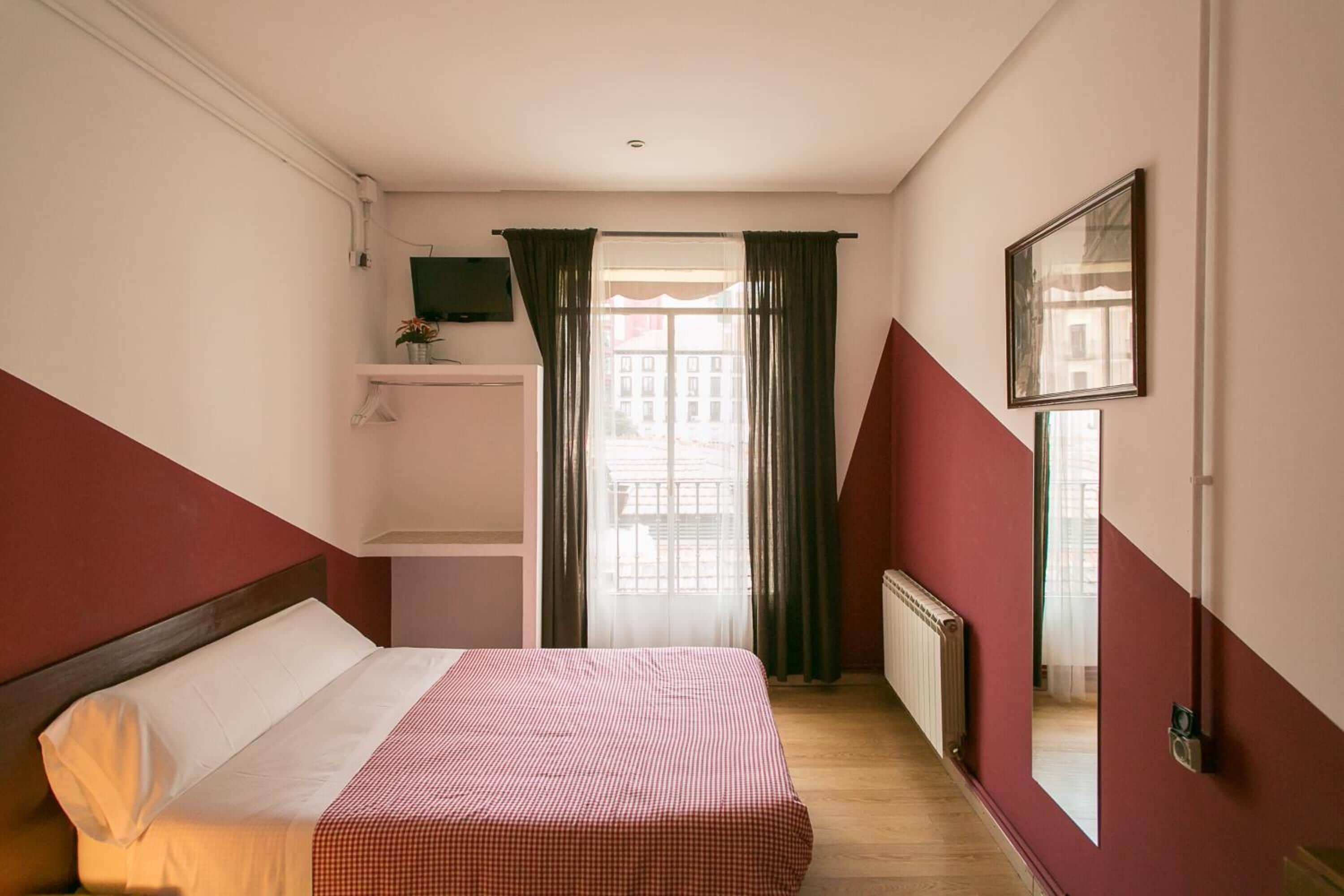 Habitación de hotel con cama, espejo, televisión, ventana con cortinas y radiador.
