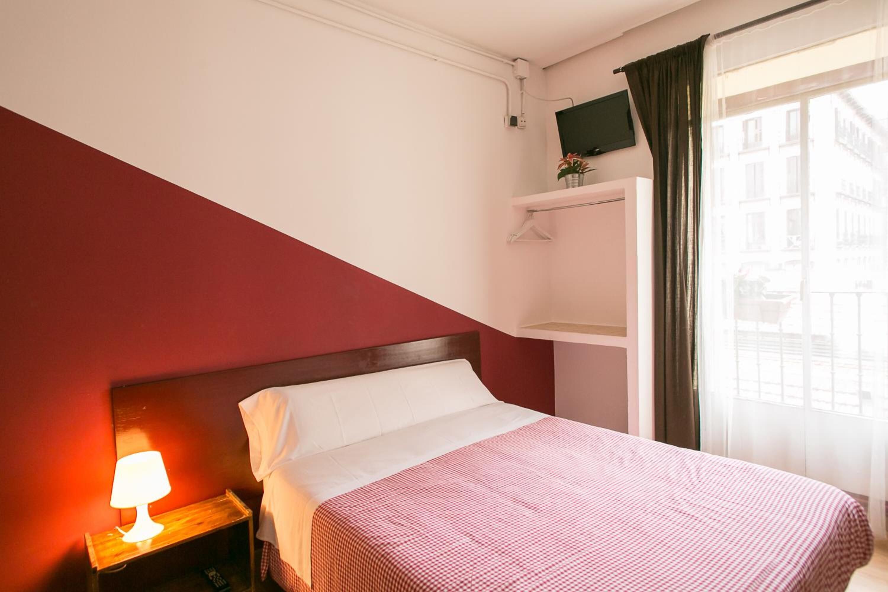 A hotel room with a bed, a nightstand with a lamp, a TV on a shelf, and a window with curtains.