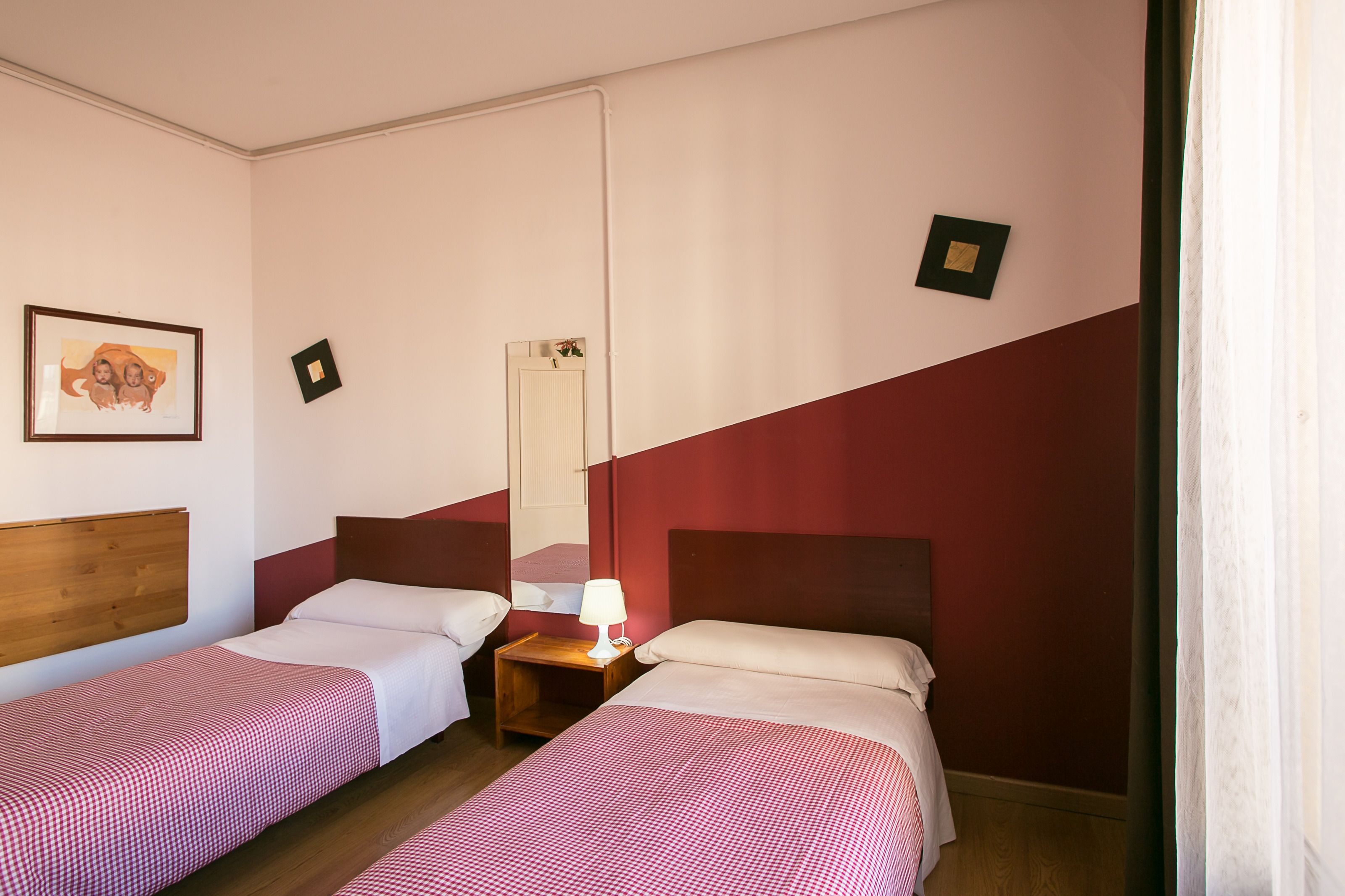 Una habitación de hotel con dos camas individuales, un cabecero de madera, una mesita de noche con lámpara, un espejo y un cuadro en la pared.