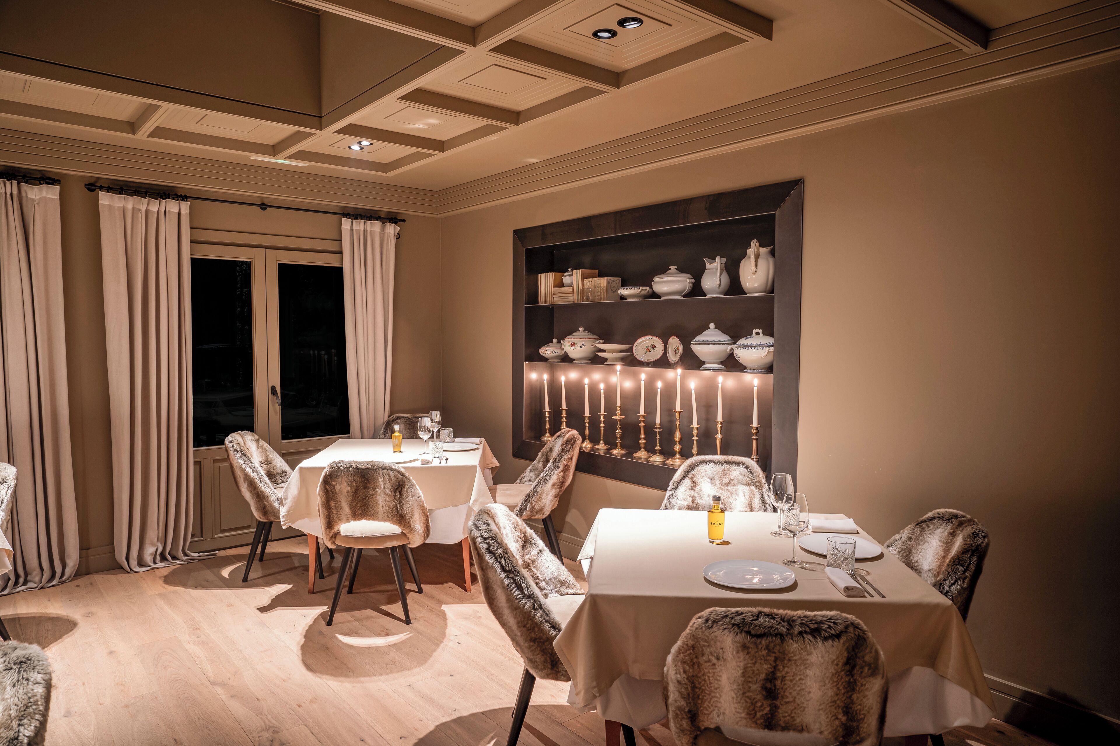 A dining area with a table set for four, surrounded by chairs with fur upholstery. A wall-mounted shelf displays various white ceramic dishes and a candelabra with candles.