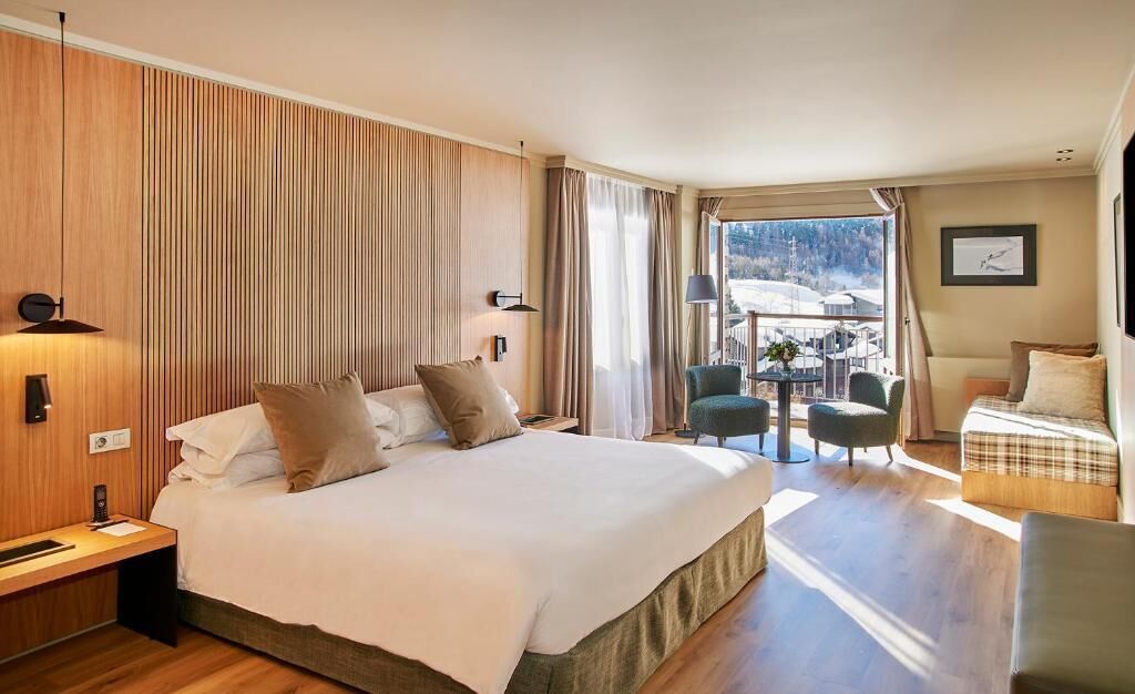 A hotel room with a large bed, a desk, a chair, a small table, and a balcony with a view.