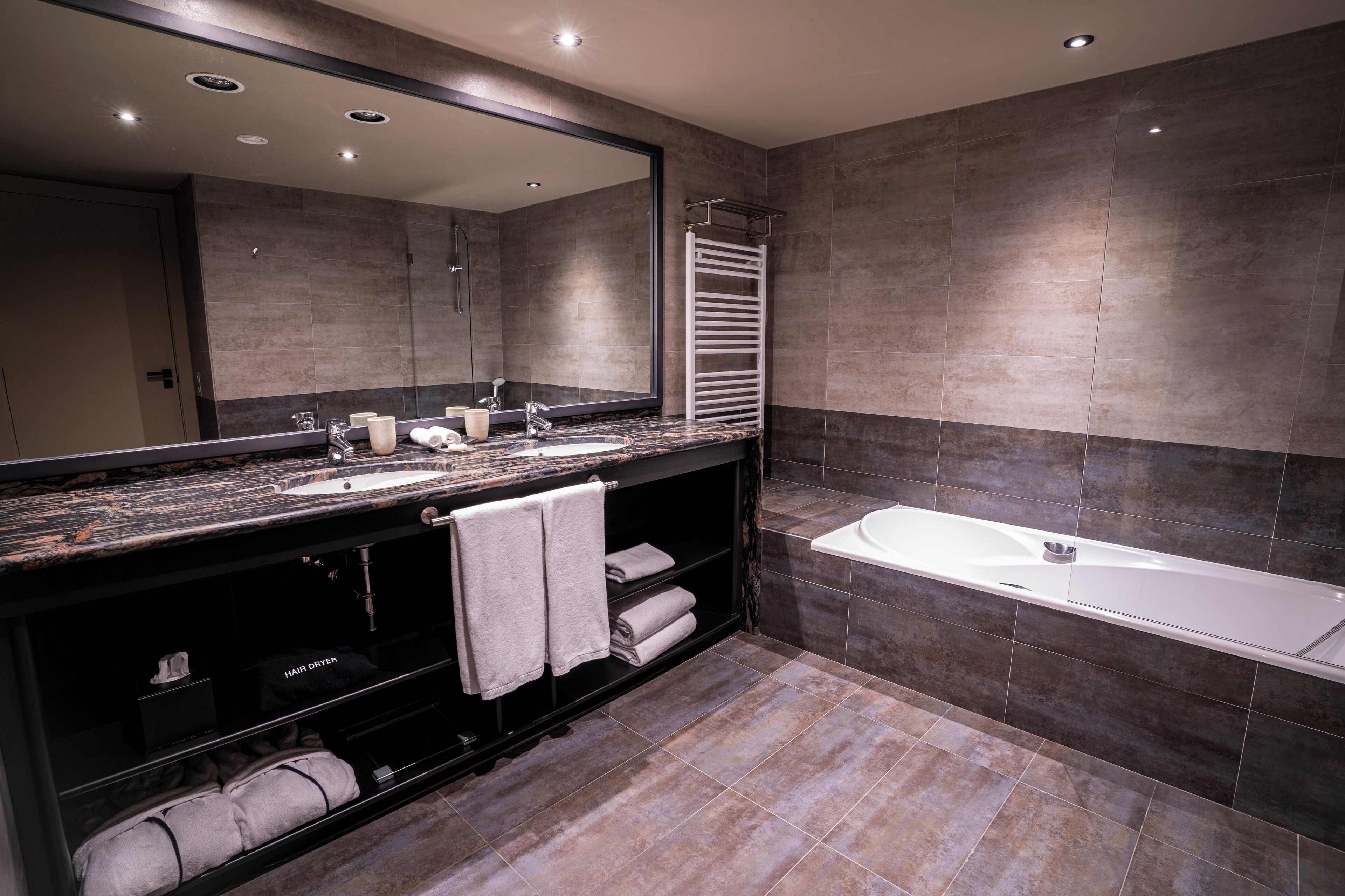 A modern bathroom with a large mirror, two sinks, a bathtub, and a towel rack.
