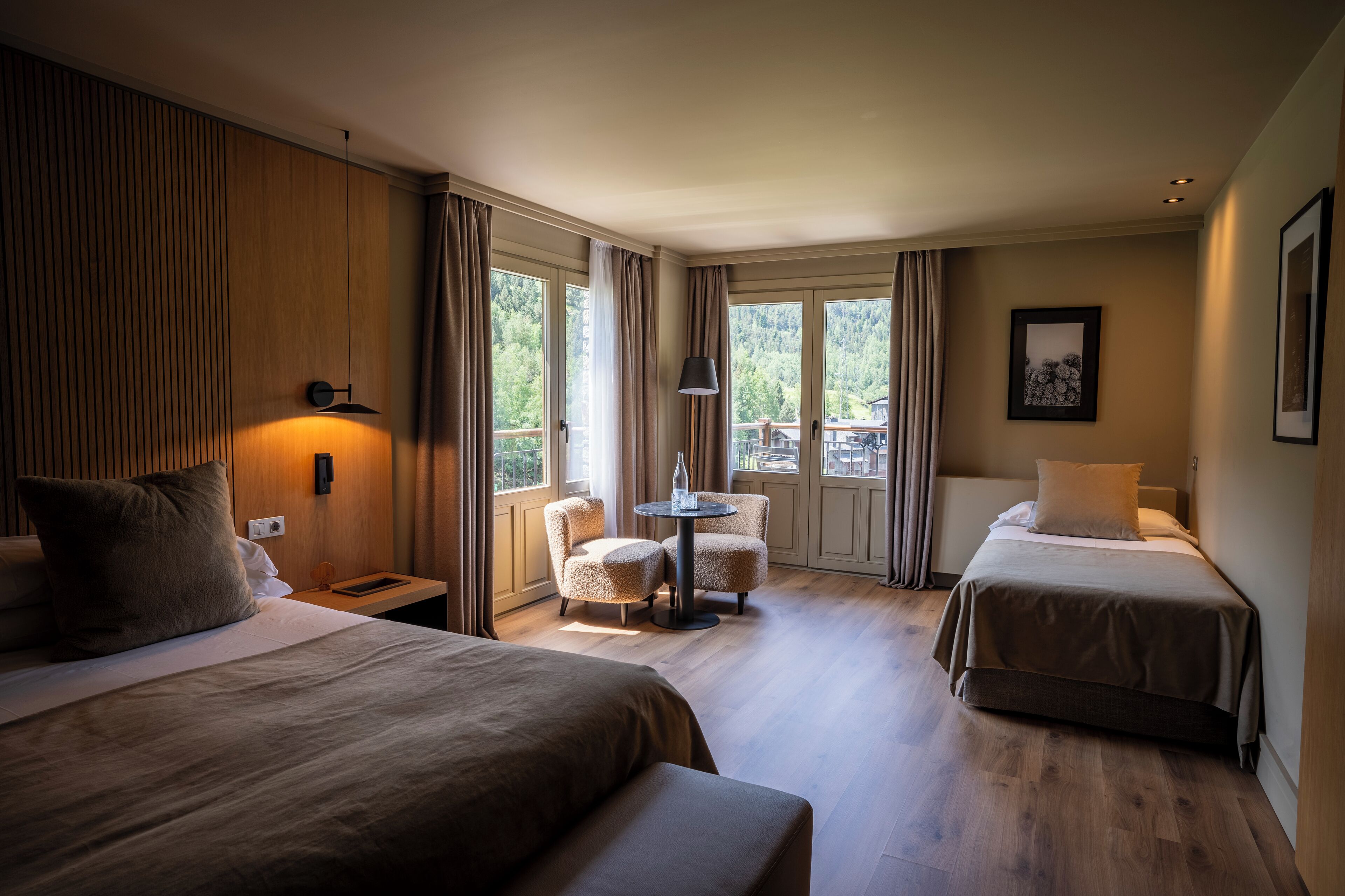 Une chambre d’hôtel avec deux lits, une petite table et une chaise. Il y a une fenêtre avec des rideaux, une lampe fixée au mur et un tableau encadré.