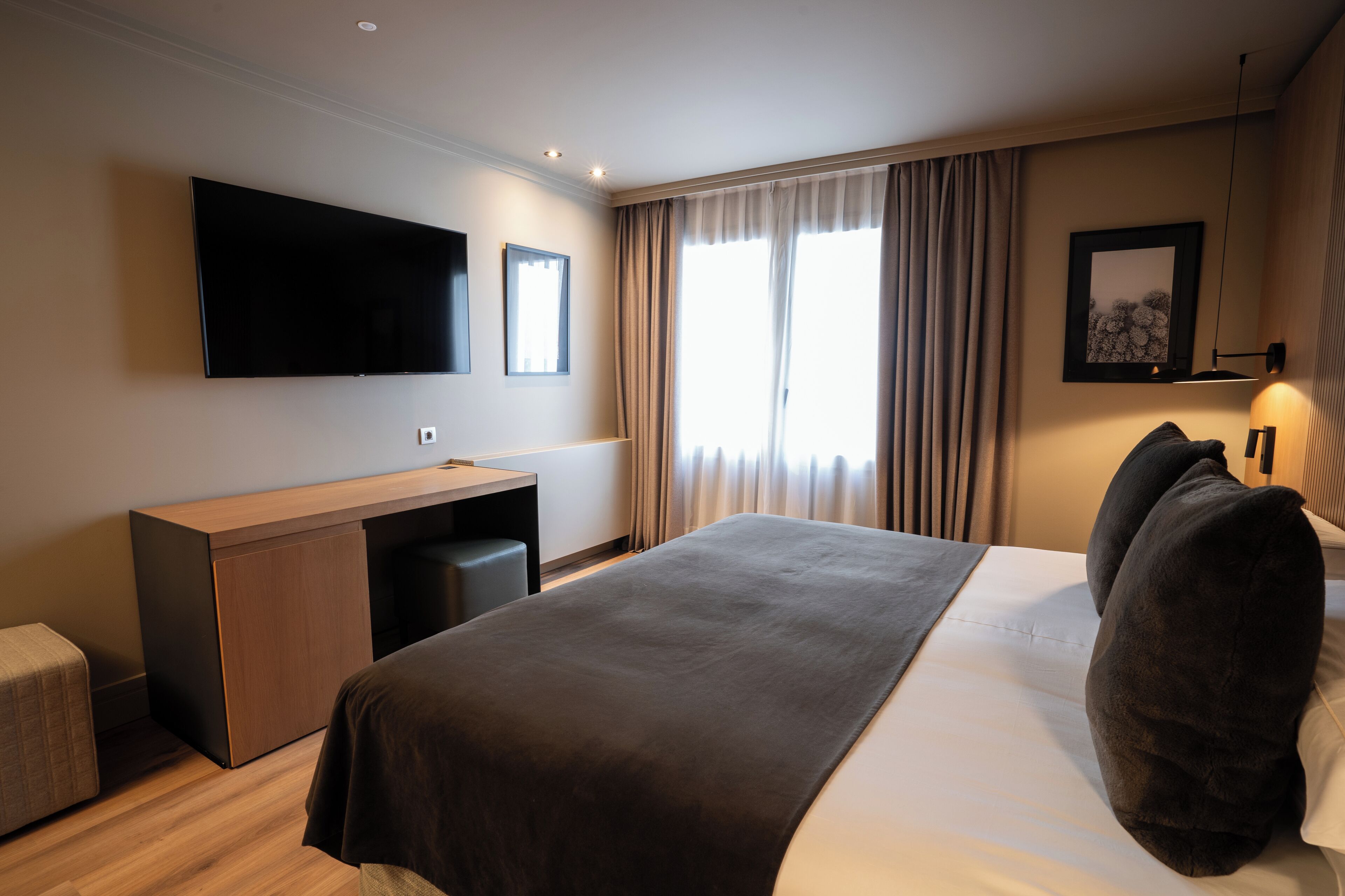 Quarto de hotel moderno com uma cama grande, uma escrivaninha, televisão de tela plana e uma janela ampla com cortinas.