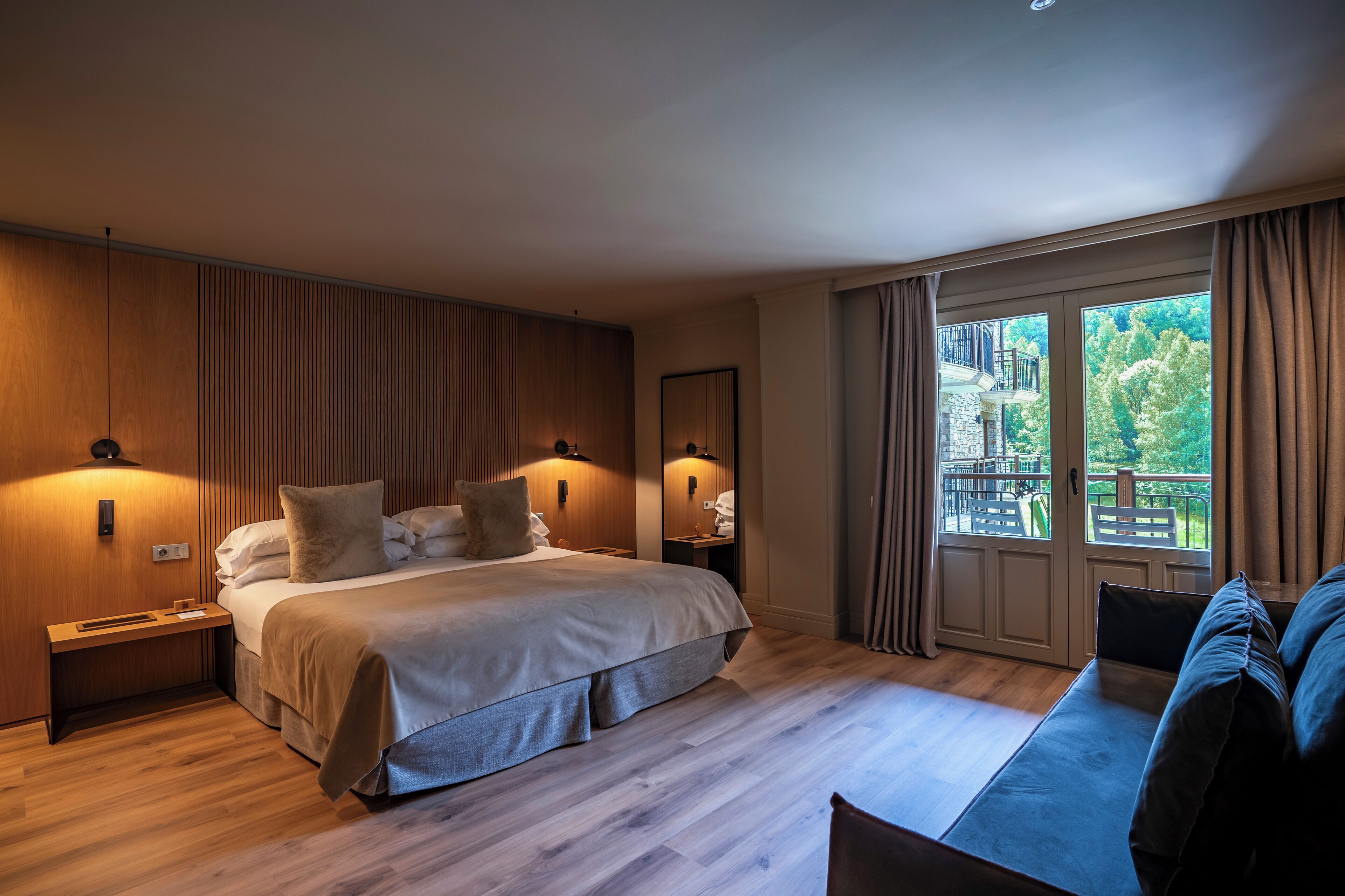 A modern hotel room with a large bed, bedside tables, a seating area, and a balcony with a view of trees.