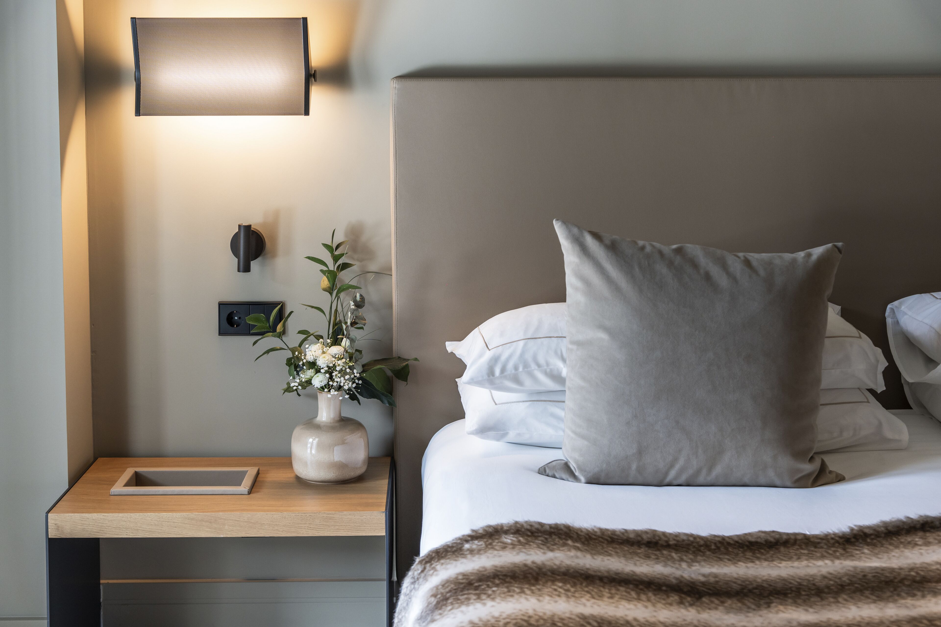 A neatly made bed with a large pillow, a bedside table with a vase of flowers, and a wall-mounted light fixture.