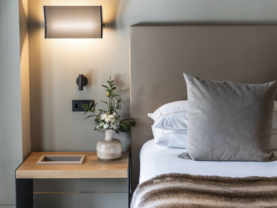 A neatly made bed with a large pillow, a bedside table with a vase of flowers, and a wall-mounted light fixture.