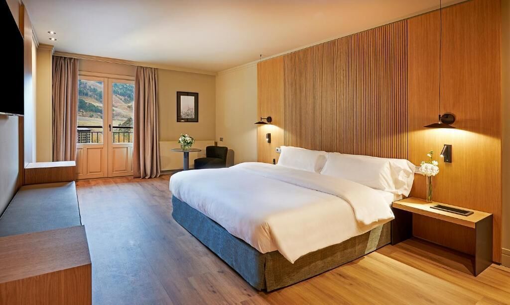 A modern hotel room with a large bed, a small desk, a chair, a TV, and a balcony with curtains.