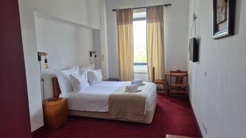 Quarto de hotel com cama, mesa de cabeceira, luminária, cadeira e janela com cortinas.