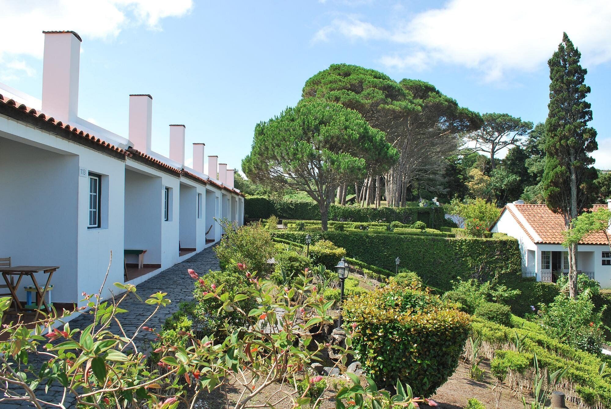 Uma fileira de edifícios brancos com telhados vermelhos, um jardim bem cuidado com sebes e árvores, e um céu claro.