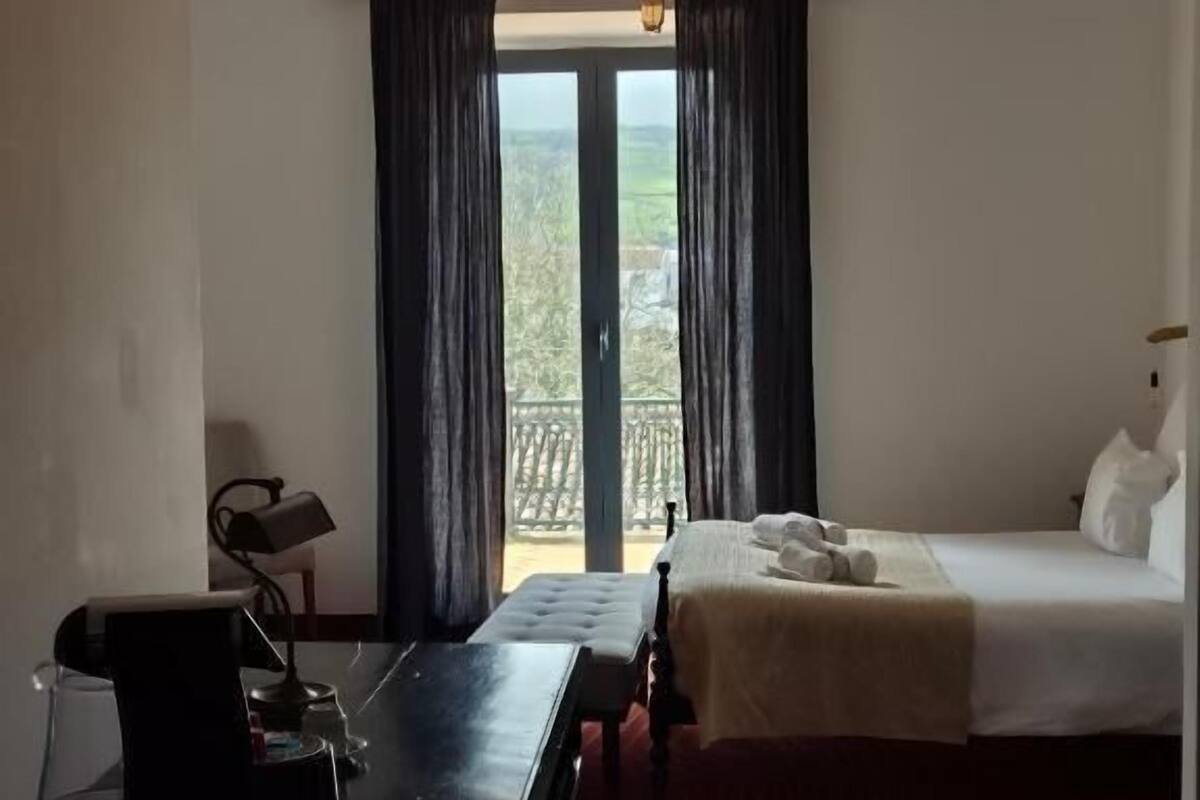 A hotel room with a bed, a desk, a chair, a lamp, and a view of the outdoors.