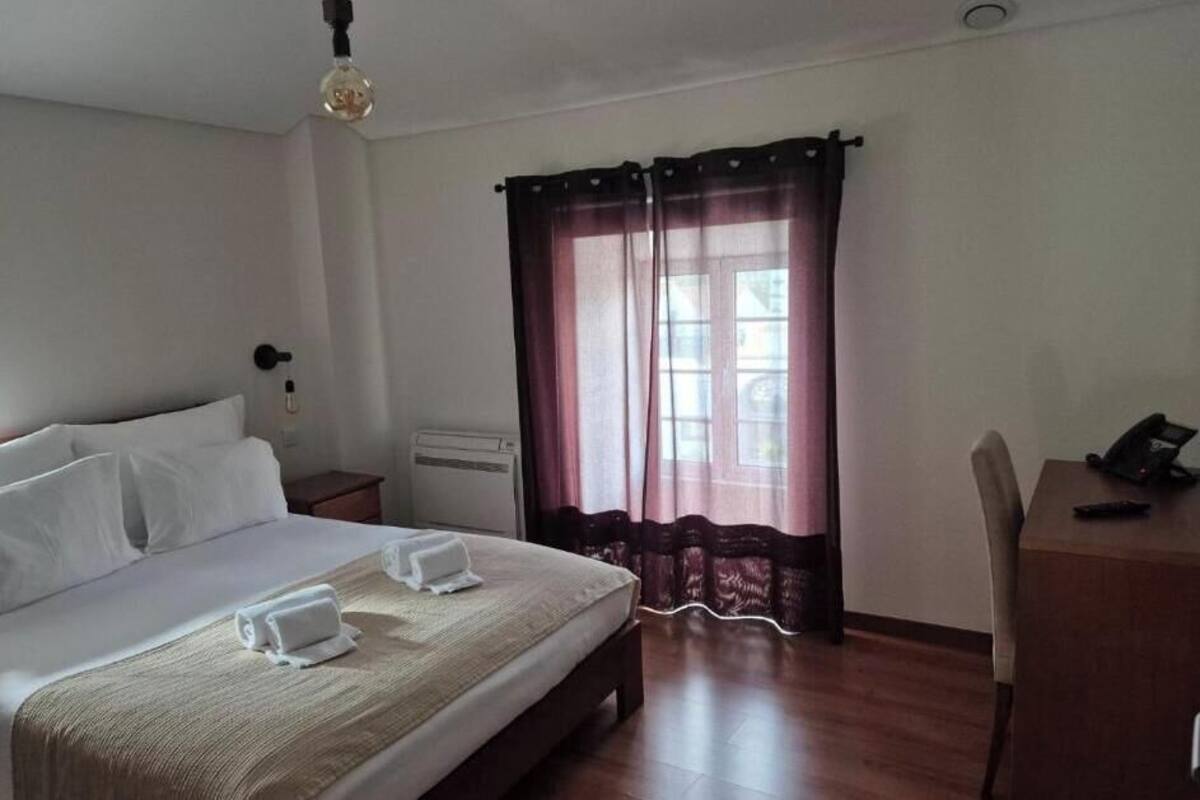A hotel room with a bed, a desk, a chair, a lamp, and a window with curtains.