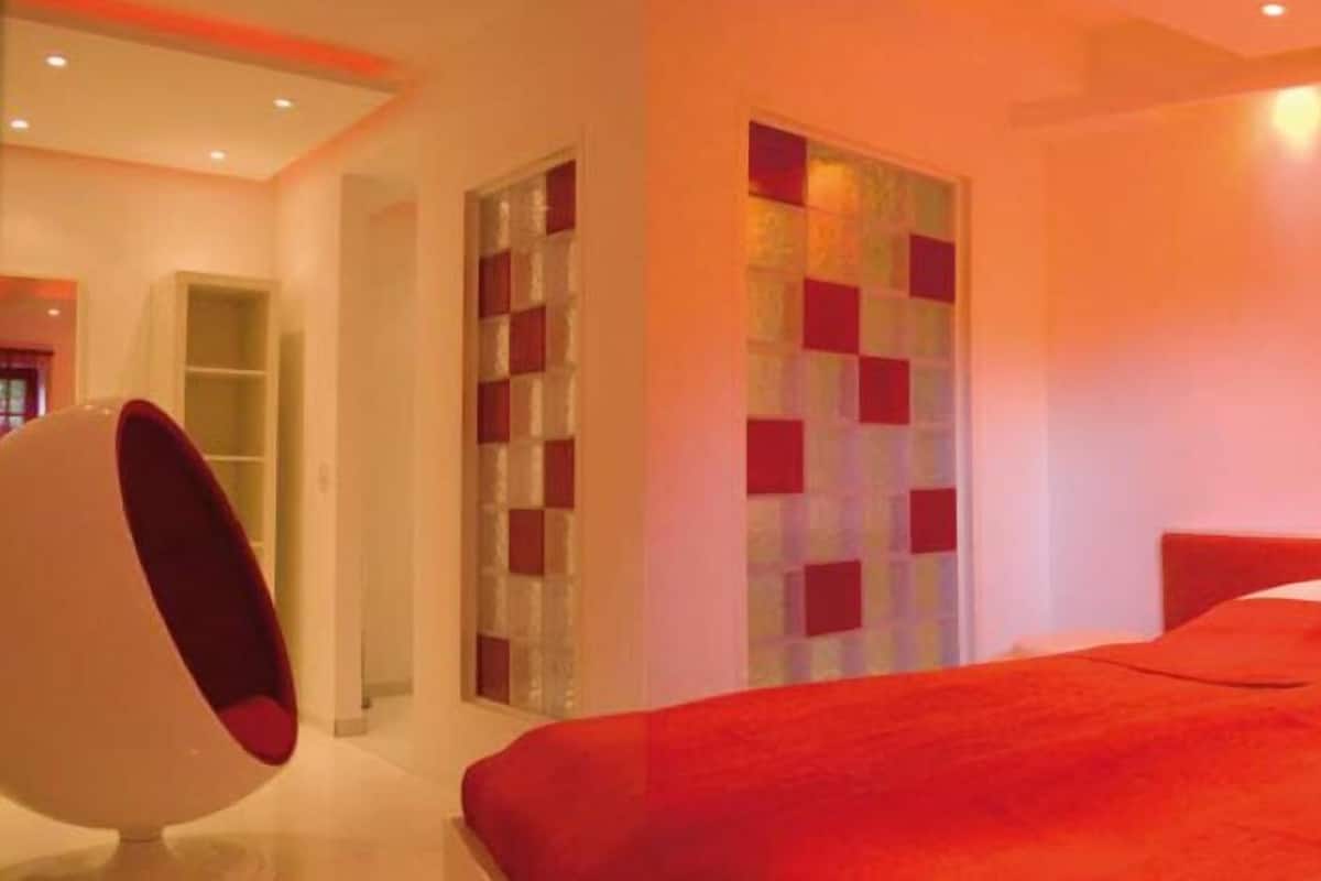 Quarto moderno com colcha vermelha, uma cadeira em formato de ovo e divisória de vidro com painéis quadrados vermelhos.