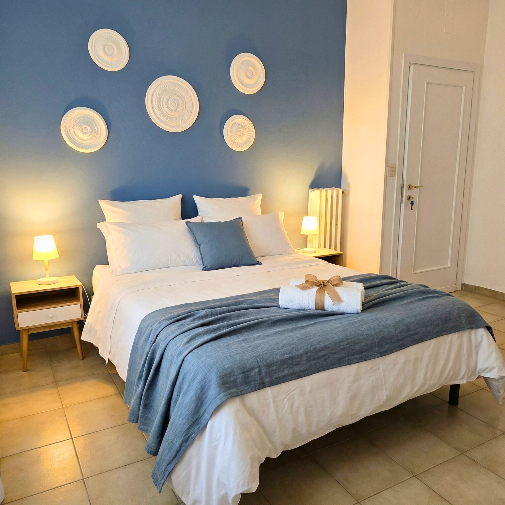 Un lit bien fait, recouvert d’une couvre-lit bleue et blanche, deux tables de chevet avec des lampes et des décorations murales.