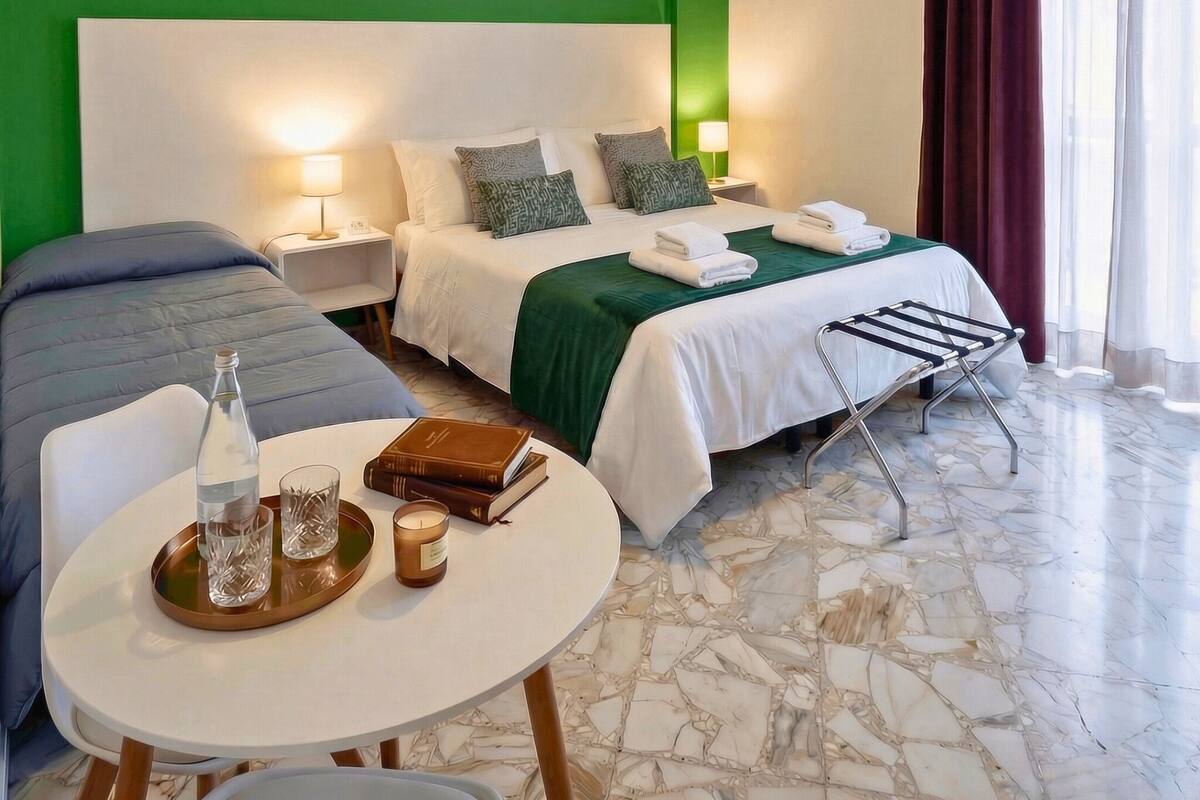 A hotel room with a green accent wall, a bed with white bedding, a bedside table with a lamp, a chair, a small table with a bottle, glasses, and books, and a marble floor.