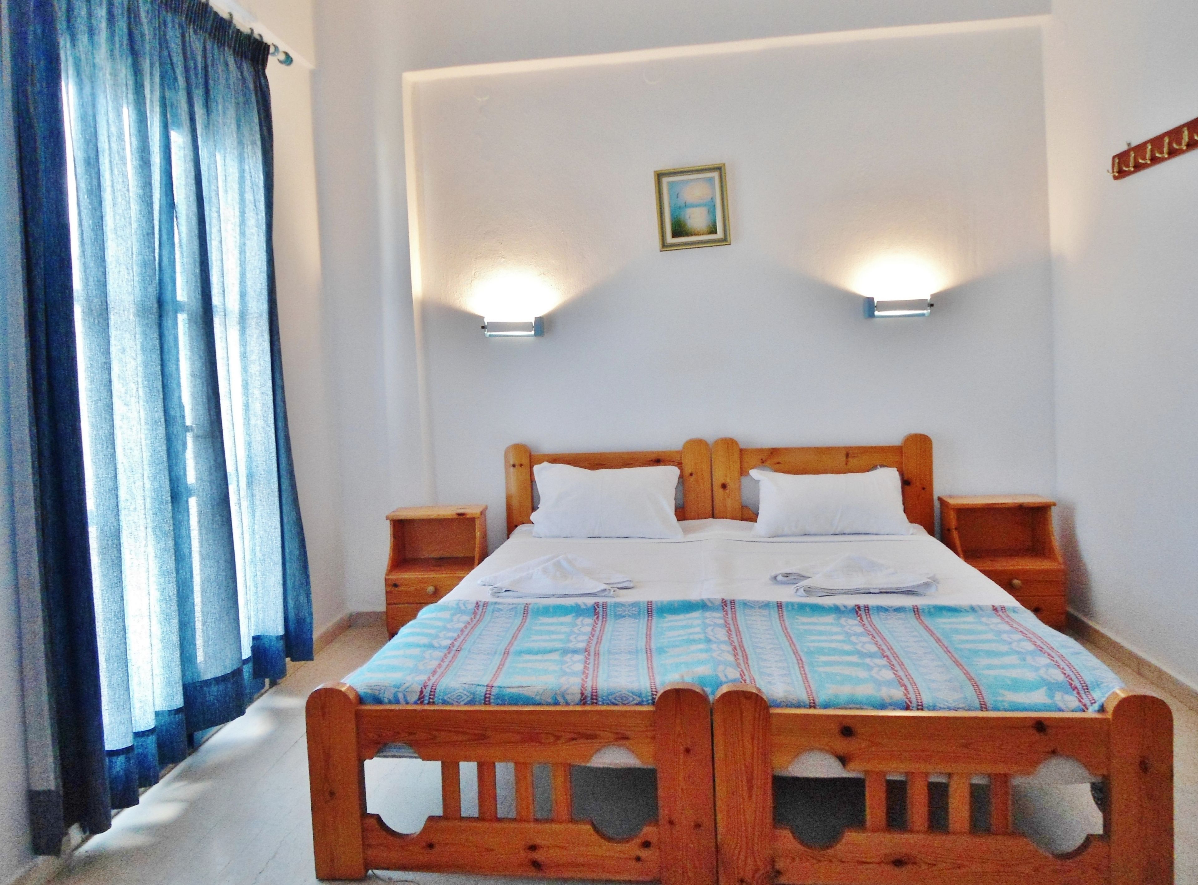 Un dormitorio con una cama de madera, dos mesitas de noche, un buró, una ventana con cortinas y luces fijadas a la pared.