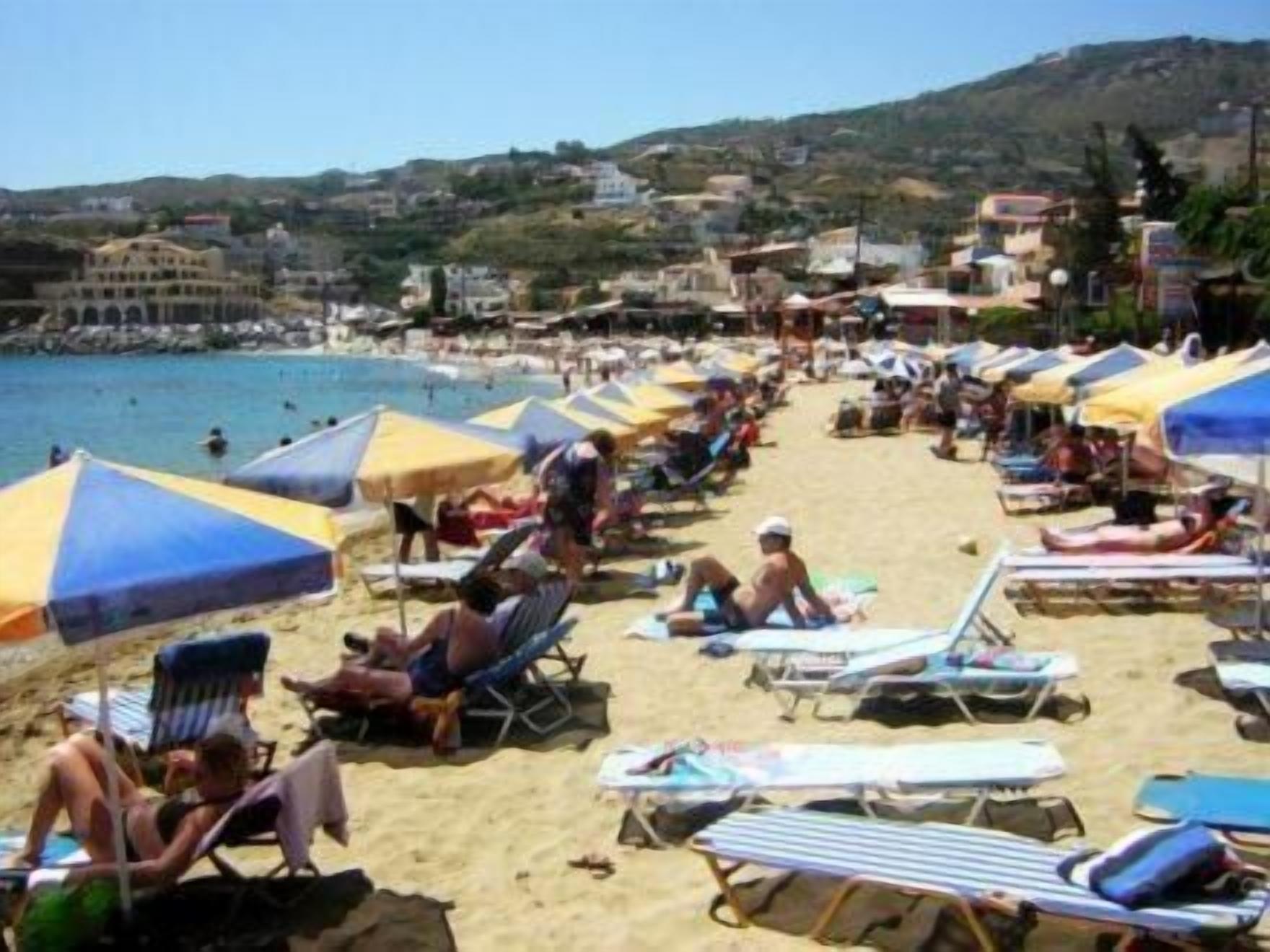 Une plage bordée de rangées de transats et de parasols, avec une vue dégagée sur la mer et des bâtiments en arrière-plan.