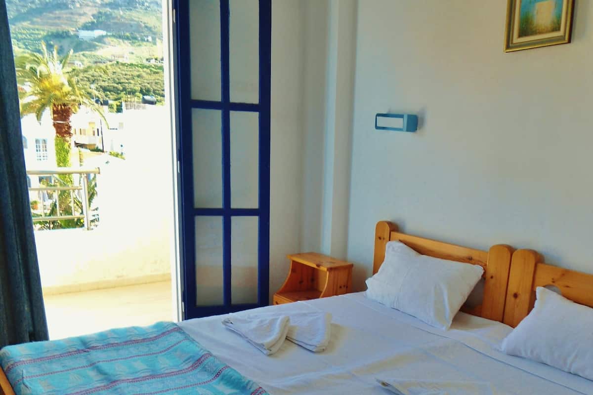 A bedroom with a wooden bed, a small wooden nightstand, a blue patterned bedspread, and a view of a balcony with a door leading outside.