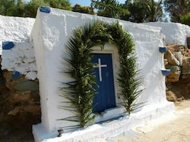 Un edificio blanco con una puerta azul, rodeado de follaje y un muro de piedra.