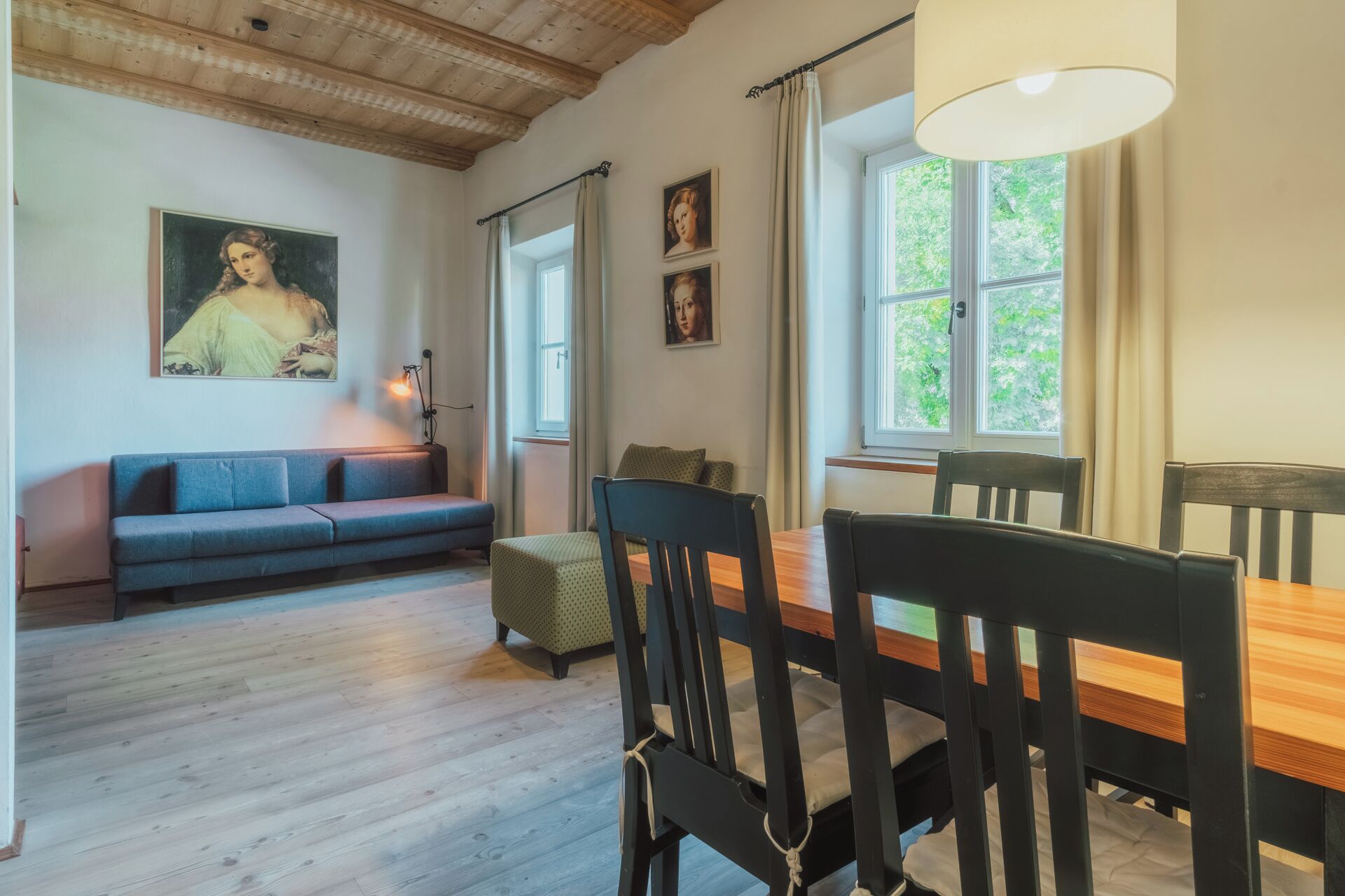 A room with a wooden ceiling, a sofa, a dining table with chairs, and a window with curtains.