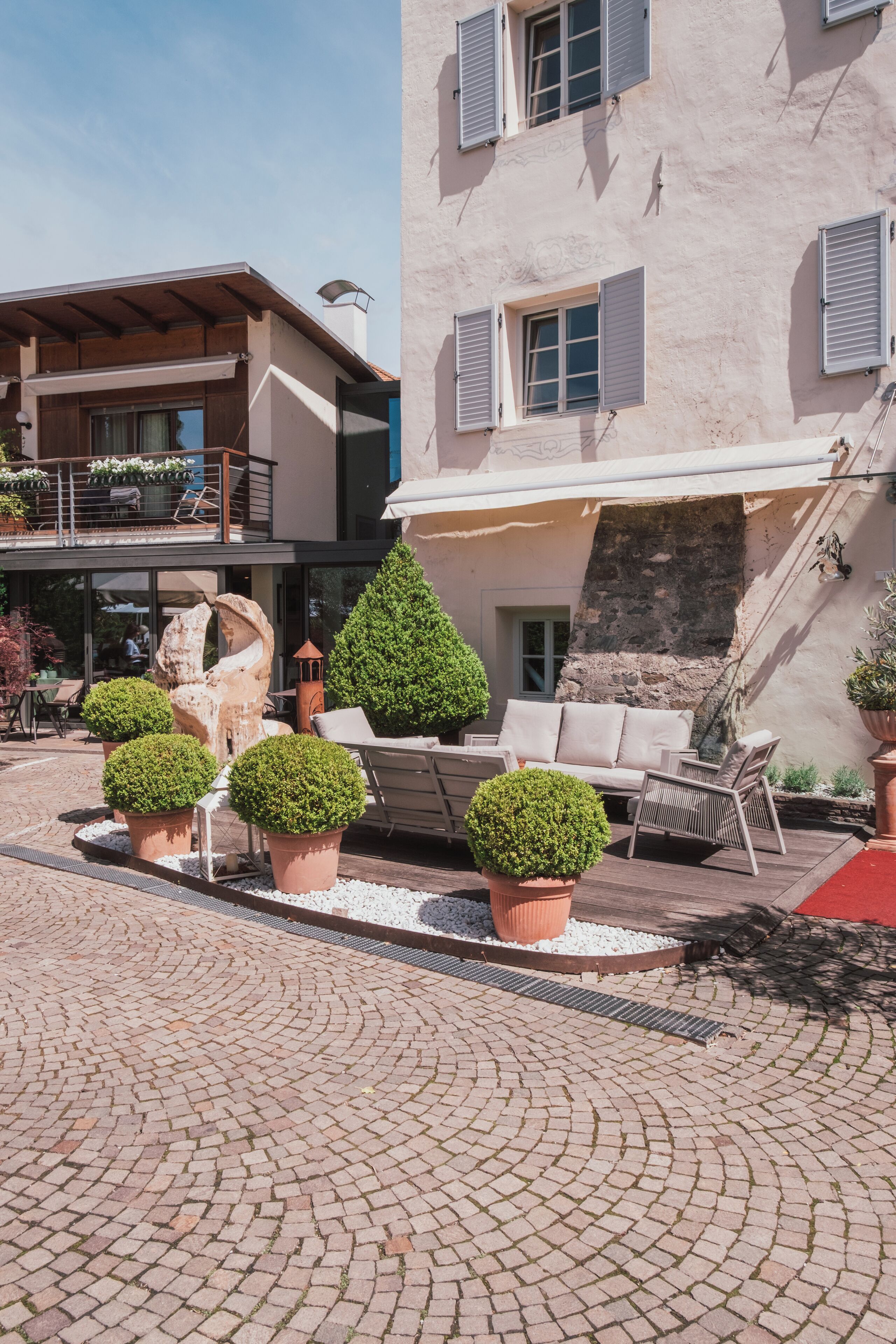 An outdoor courtyard with a seating area, potted plants, and a building with a stone facade and shutters.