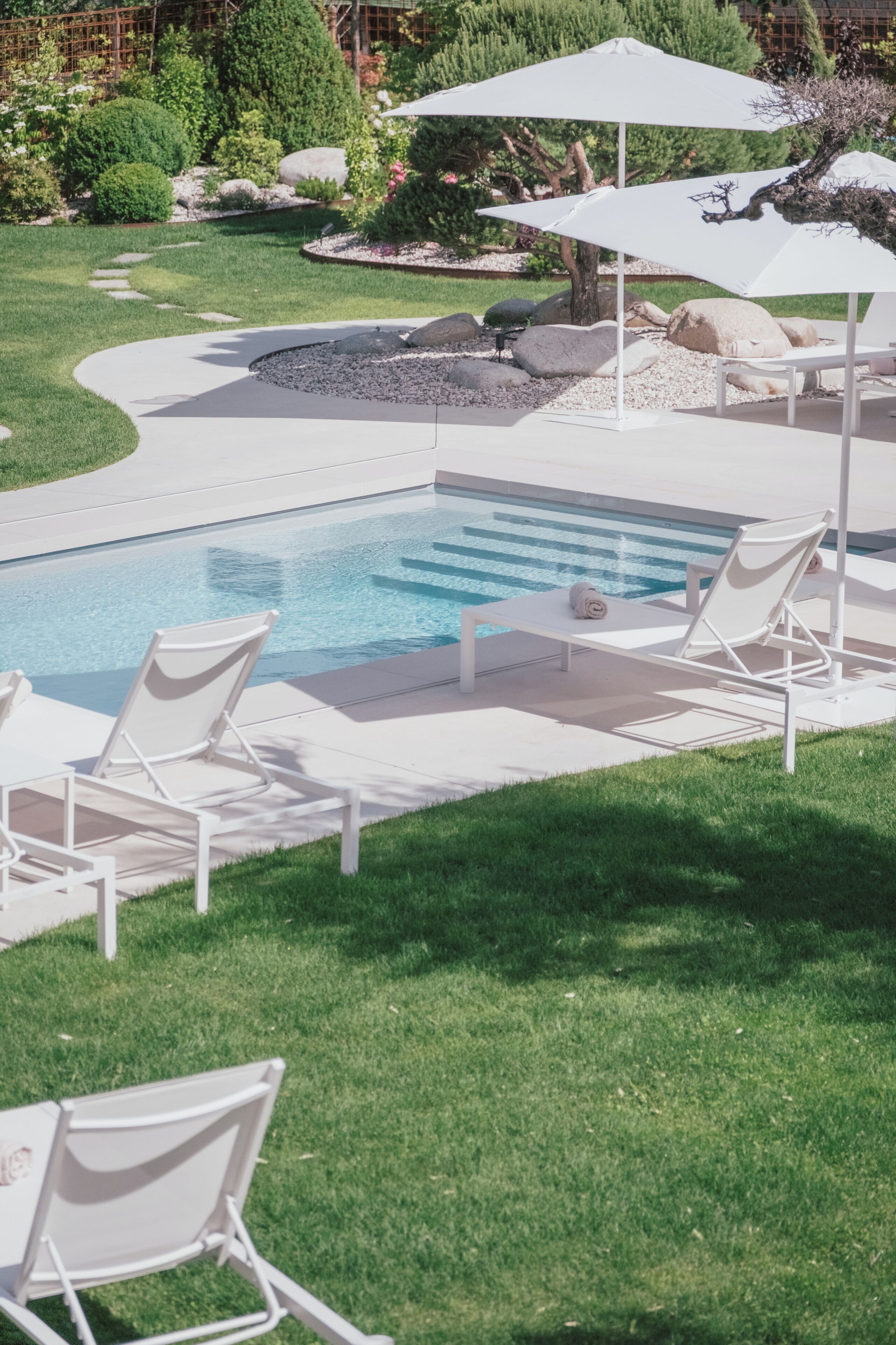 A swimming pool with white lounge chairs and umbrellas, surrounded by a well-maintained lawn and rocks.