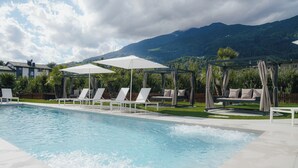 A swimming pool with lounge chairs and umbrellas, surrounded by a wooden fence and greenery, with mountains in the background. - Hotel Traube (Bressanone)