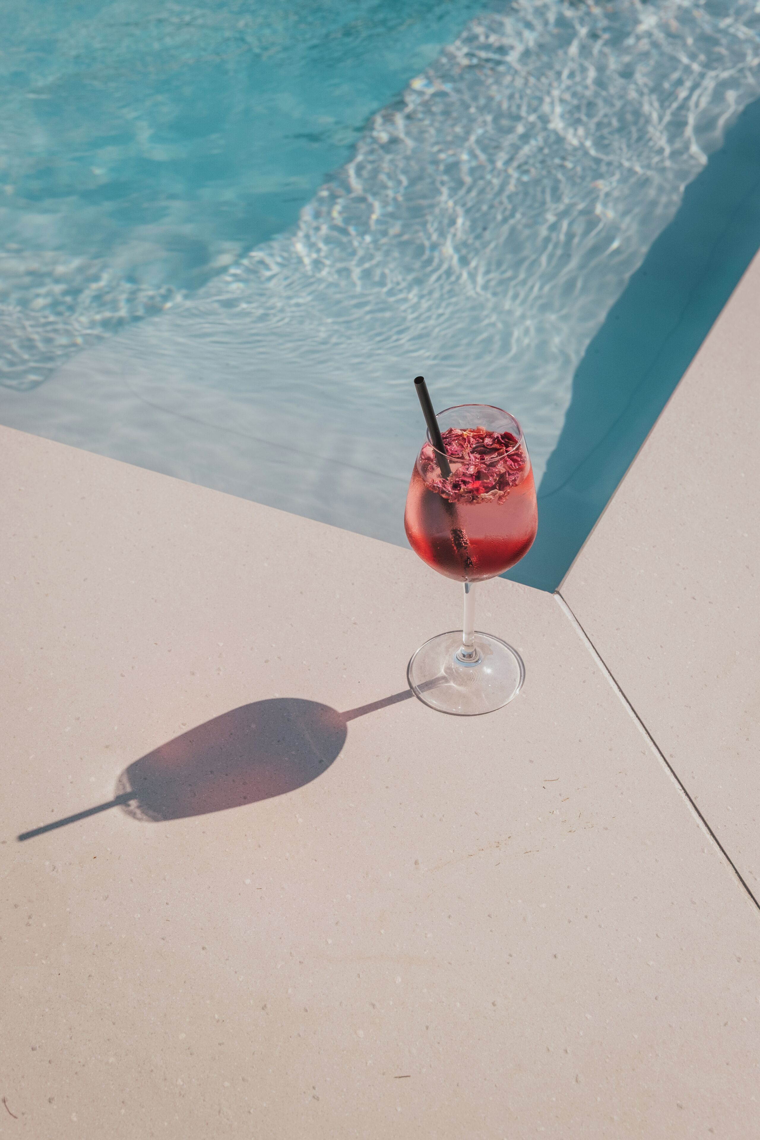 A glass of red drink with a straw on a poolside with clear blue water.