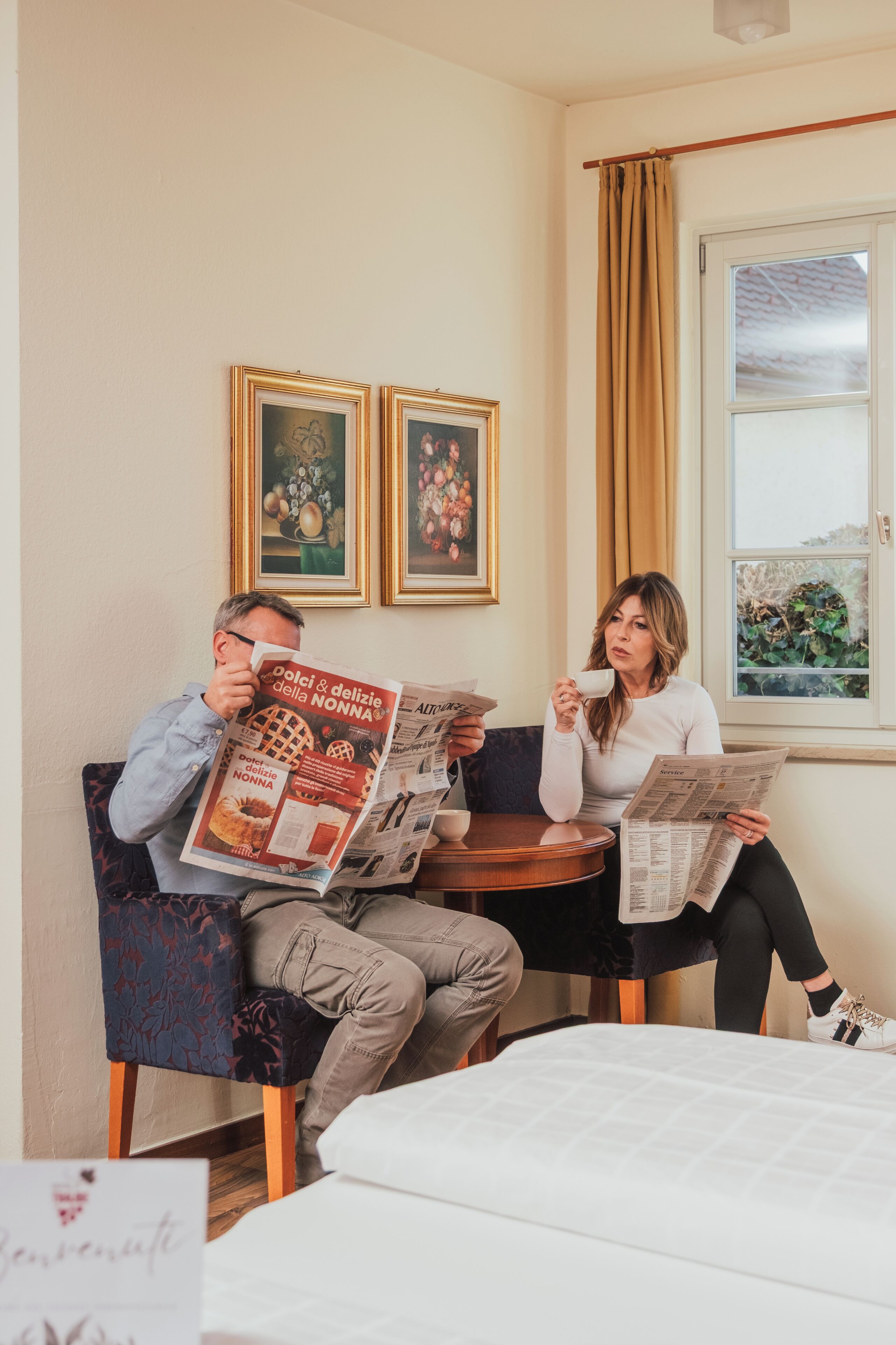 Two people seated at a round table in a room with a bed, a window with curtains, and framed pictures on the wall.