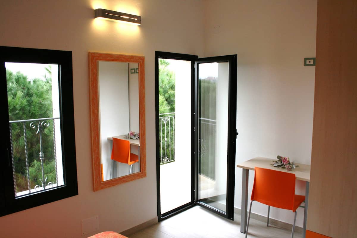 Habitació amb un llit, un escriptori amb una cadira taronja, un mirall i una porta corredissa de vidre que dona a un balcó amb vistes a vegetació.